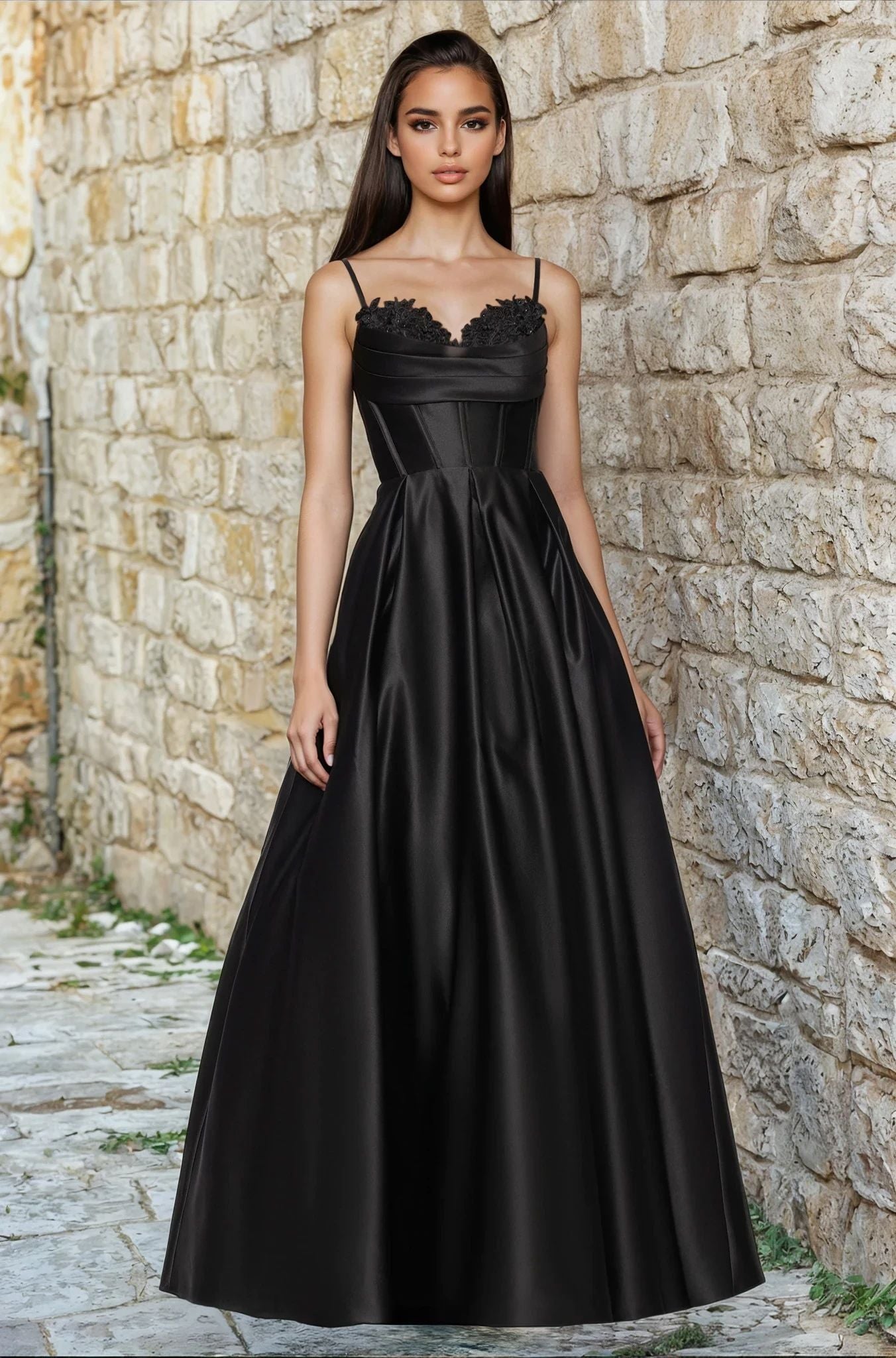 A woman stands outdoors against a stone wall, wearing the Blondie Nites 5453BN long prom dress—a sleeveless black gown with lace applique on the bodice and thin straps.